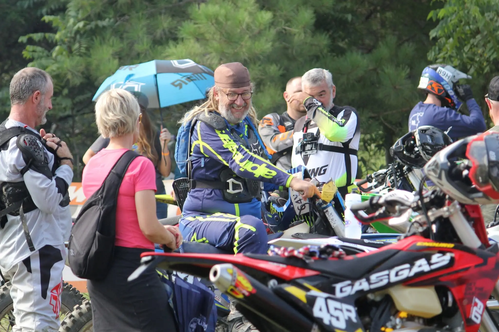 Riders and family members gathered around the bikes before a race, one rider laughing, under the shade of an umbrella